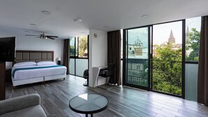Une chambre d’hôtel moderne avec un grand lit, une table ronde, un ventilateur de plafond et une vue sur le paysage urbain, visible à travers de grandes fenêtres.
