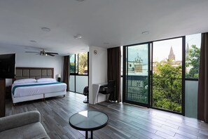 Quarto de hotel moderno com uma cama grande, uma mesa redonda, ventoinha no teto e vista para a cidade através de janelas amplas.