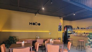 L'intérieur d'un restaurant, avec des tables et des chaises, un comptoir où est affiché le menu, et un mur sur lequel on peut lire le mot « MODO ».