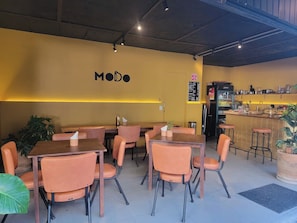Interior de restaurante com mesas, cadeiras, um balcão com um menu e uma parede com a palavra 'MODO'.
