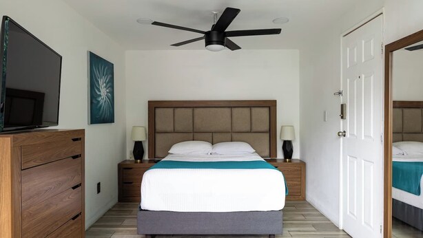 Une chambre à coucher comprenant un lit, des tables de chevet, un ventilateur de plafond, un téléviseur et une commode en bois.