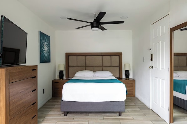 Quarto com cama, mesas de cabeceira, ventoinha no teto, televisão e cômoda de madeira.