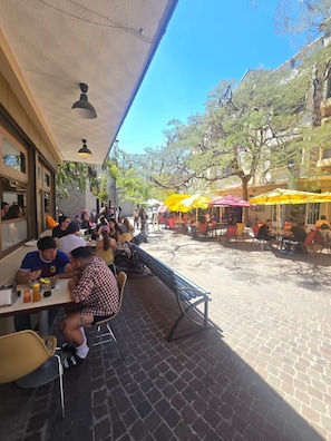 Um café ao ar livre com mesas e cadeiras, rodeado por árvores e edifícios.