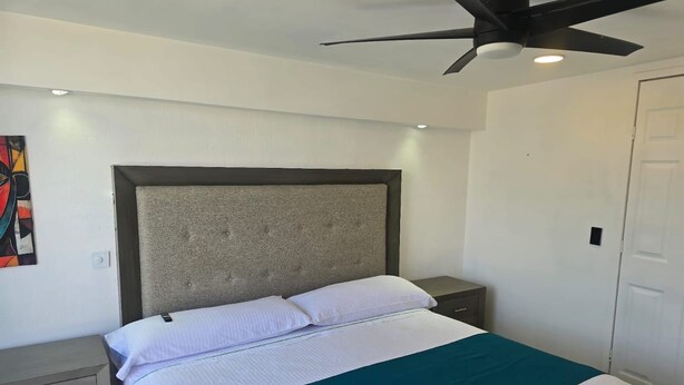 Une chambre à coucher avec un lit, un ventilateur de plafond, une table de chevet, une commode et une porte.