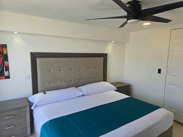 Quarto com cama, ventoinha no teto, mesa de cabeceira, cômoda e porta.