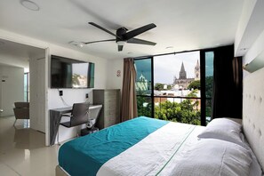 Quarto de hotel com cama, escrivaninha e vista para uma catedral através de amplas janelas.