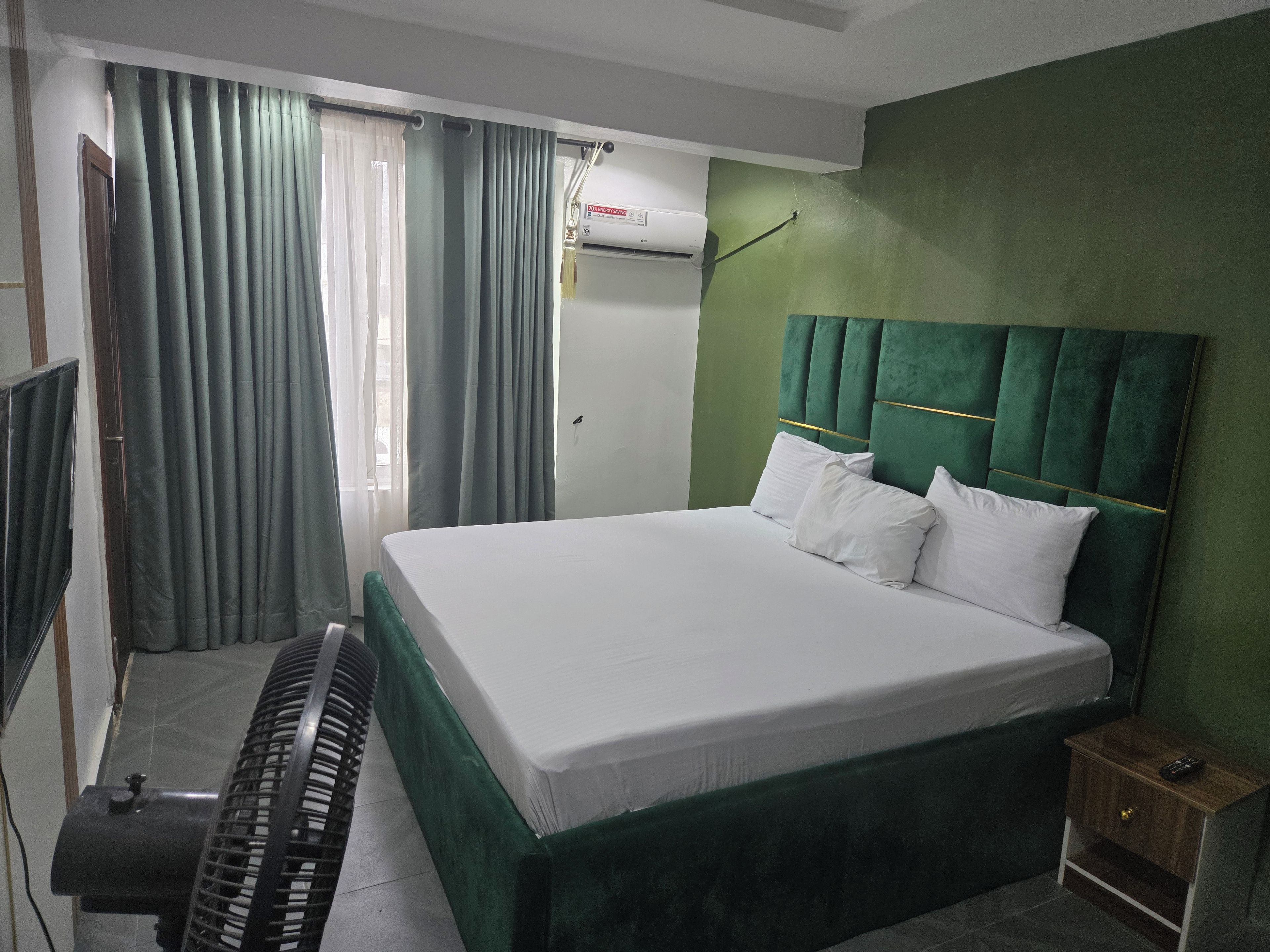 Habitació d'hotel amb un llit, una tauleta de nit, un ventilador i una finestra amb cortines.