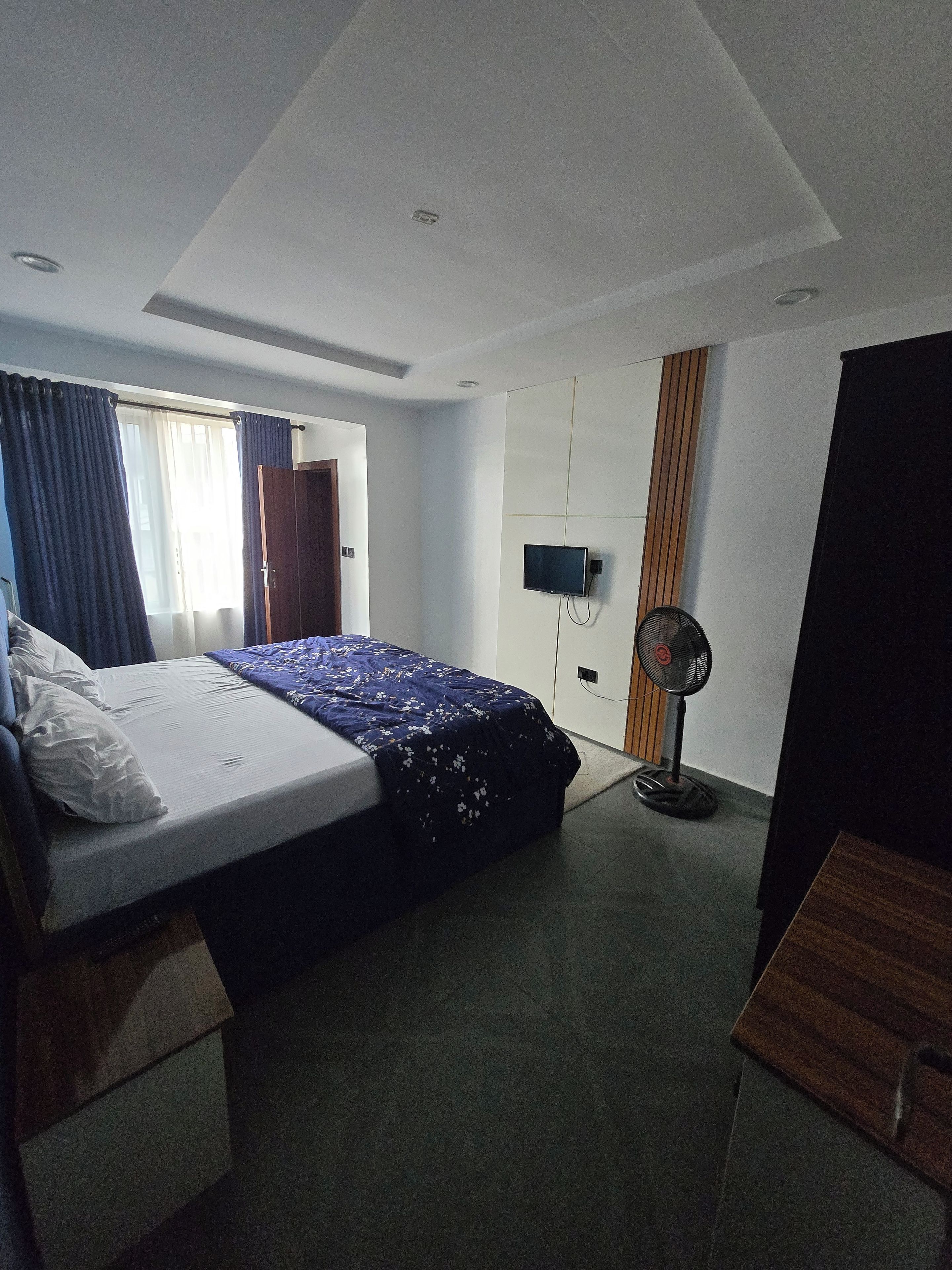 A hotel room with a bed, a television, a standing fan, and a wooden wardrobe.