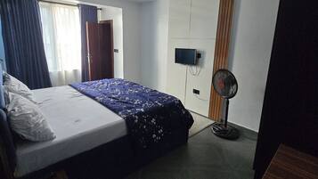 A hotel room with a bed, a television, a standing fan, and a wooden wardrobe.