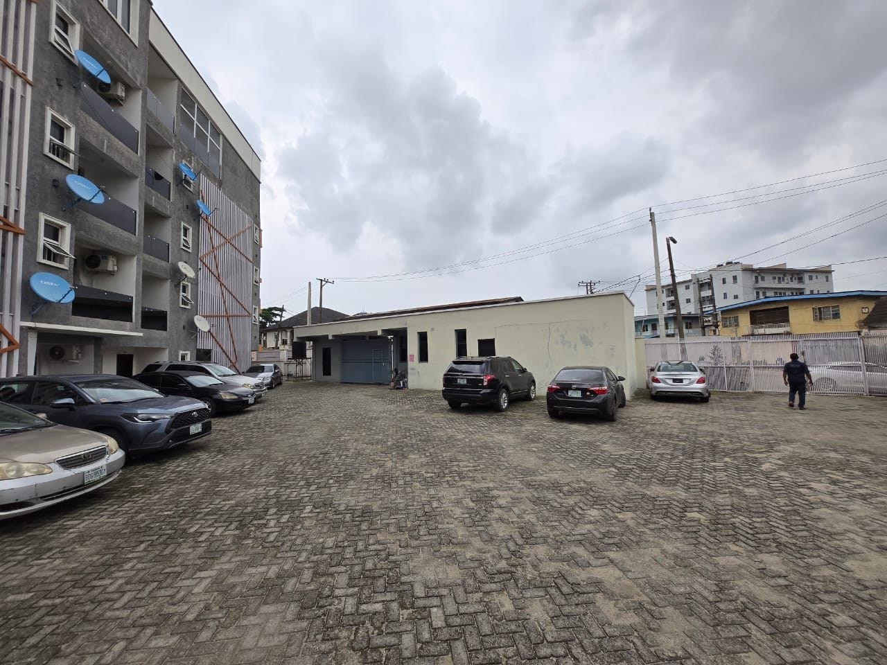 Una zona aparcament amb diversos cotxes, un edifici amb garatge i una persona caminant.