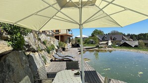 Una terraza de madera con una mesa y sillas, un paraguas grande, y vista a un estanque con casas al fondo.