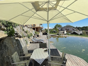 Una terraza de madera con una mesa y sillas, un paraguas grande, y vista a un estanque con casas al fondo.