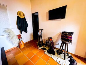 A room with a tiled floor, a television mounted on the wall, a hat, a jacket, and a small stool with items on it. - PARADOR LA MESA REDONDA (Los Santos)