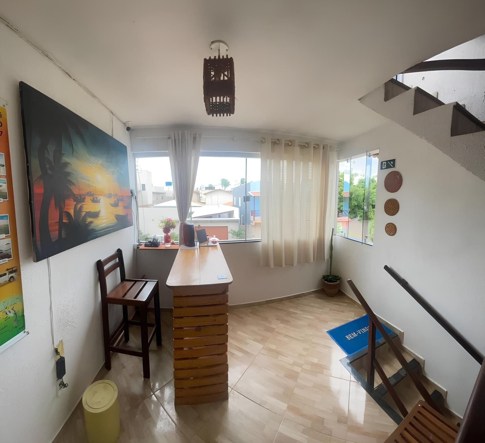 A room with a wooden table, chairs, a window with curtains, a painting on the wall, and a staircase.