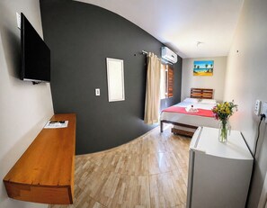 A compact hotel room with a bed, a desk, a TV, and an air conditioning unit. - Vila Cactos Jeri (Jijoca de Jericoacoara)