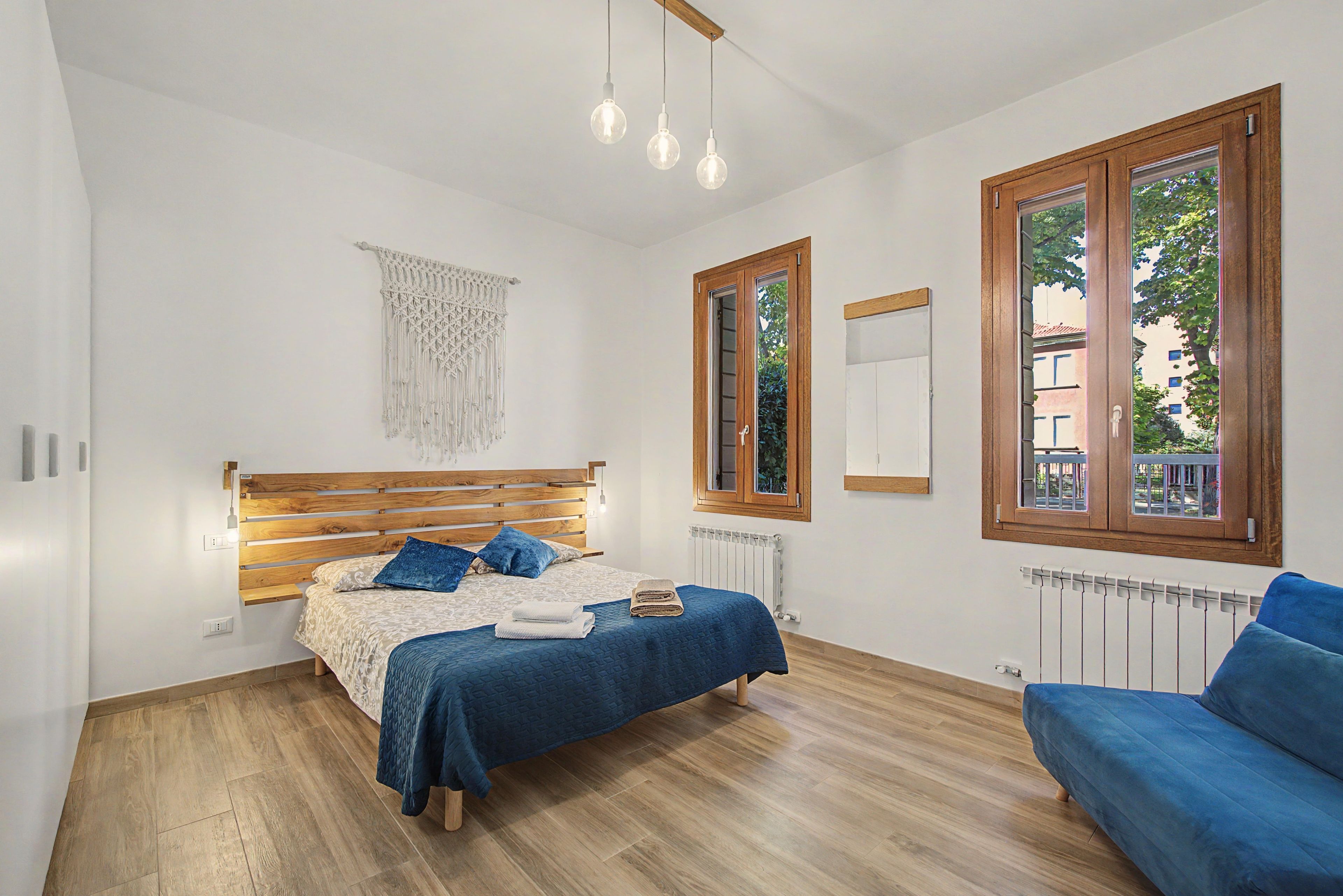 A bedroom with a wooden bed, a blue sofa, a macrame wall hanging, and a window with a view of buildings.