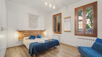 A bedroom with a wooden bed, a blue sofa, a macrame wall hanging, and a window with a view of buildings.