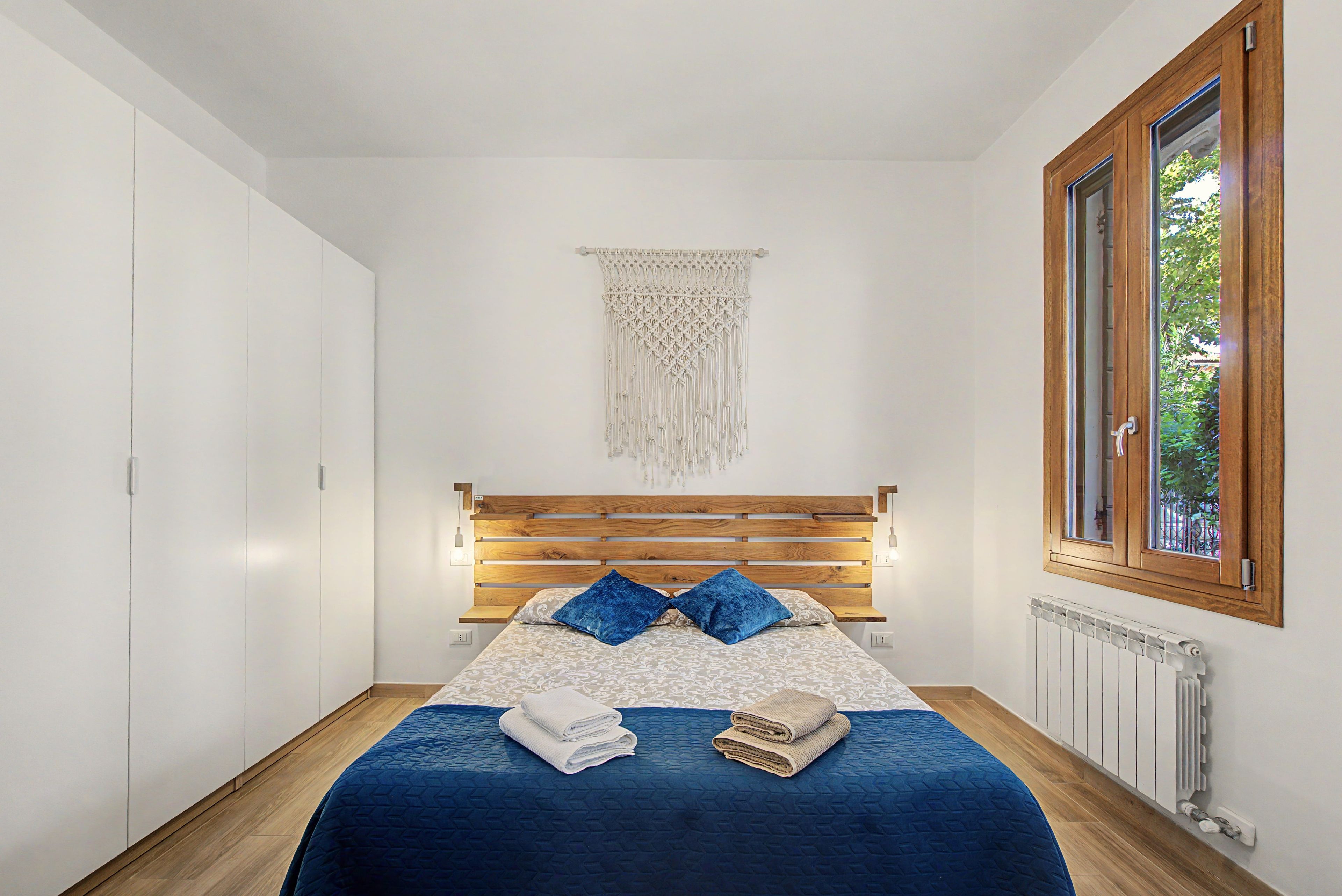 Ein Schlafzimmer mit einem Bett aus Holz, einer blauen Bettdecke, zwei Kissen, einer Wanddekoration aus Makramee und einem Fenster mit Blick auf Grün.