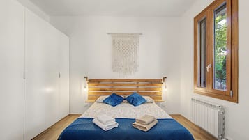 Ein Schlafzimmer mit einem Bett aus Holz, einer blauen Bettdecke, zwei Kissen, einer Wanddekoration aus Makramee und einem Fenster mit Blick auf Grün.