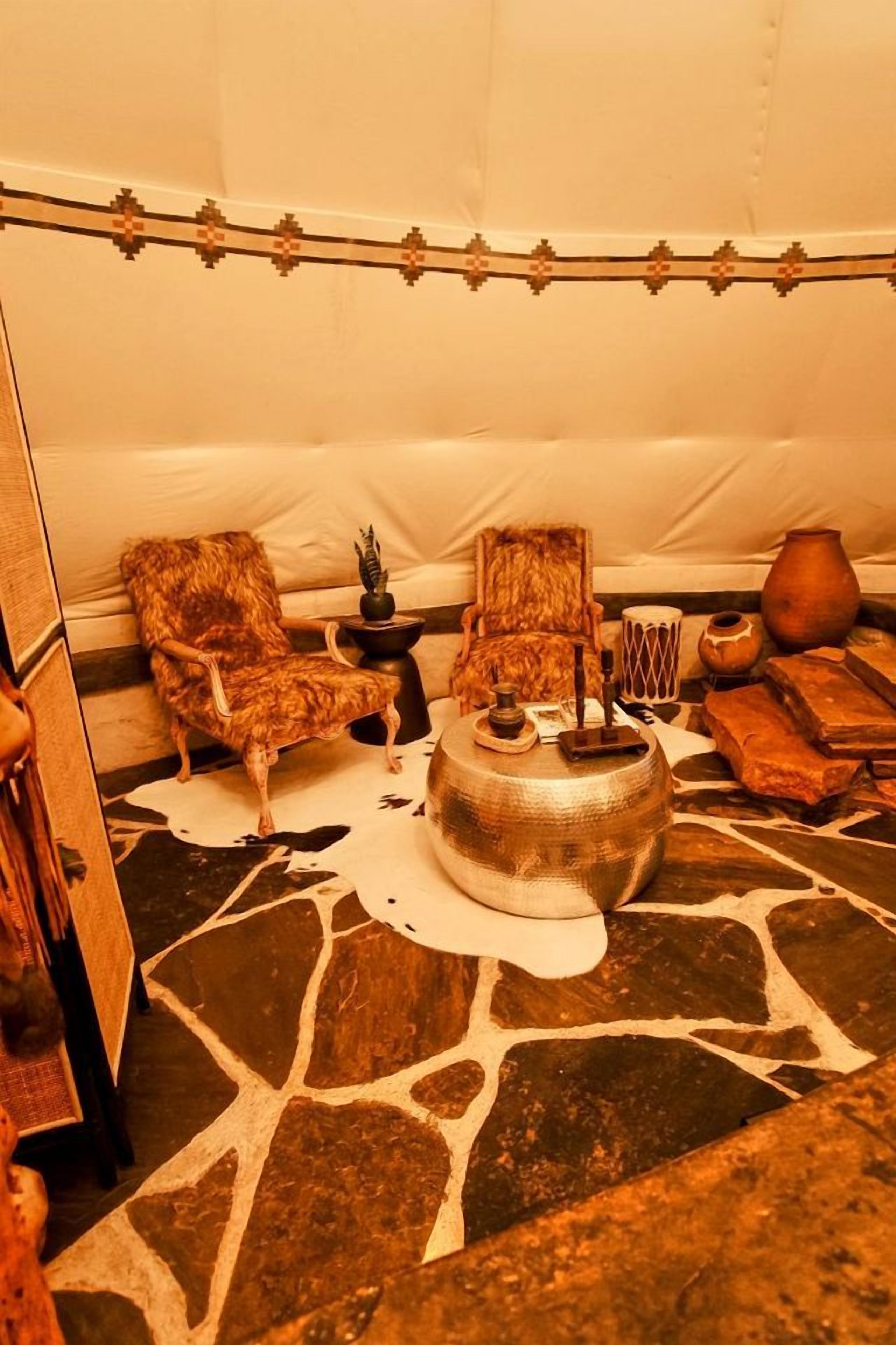 A tent interior with a stone floor, two chairs, a central table with a lamp, and various decorative items.