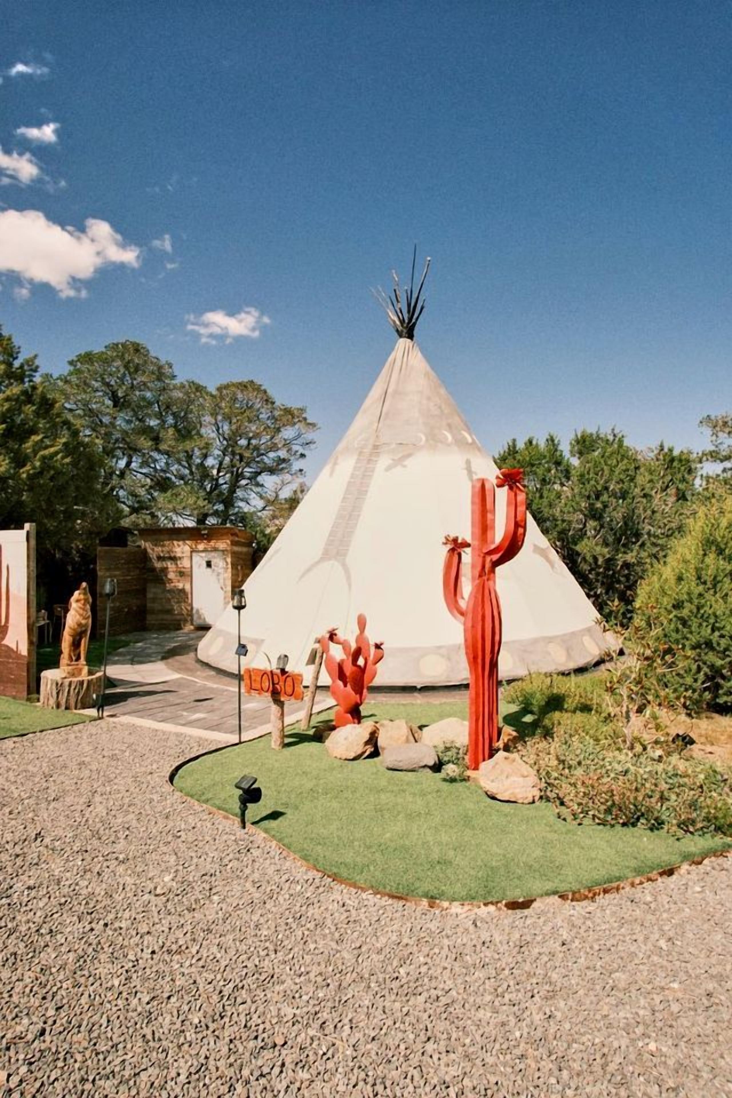 A tipi with a grassy area in front, a red sculpture, and a stone structure.