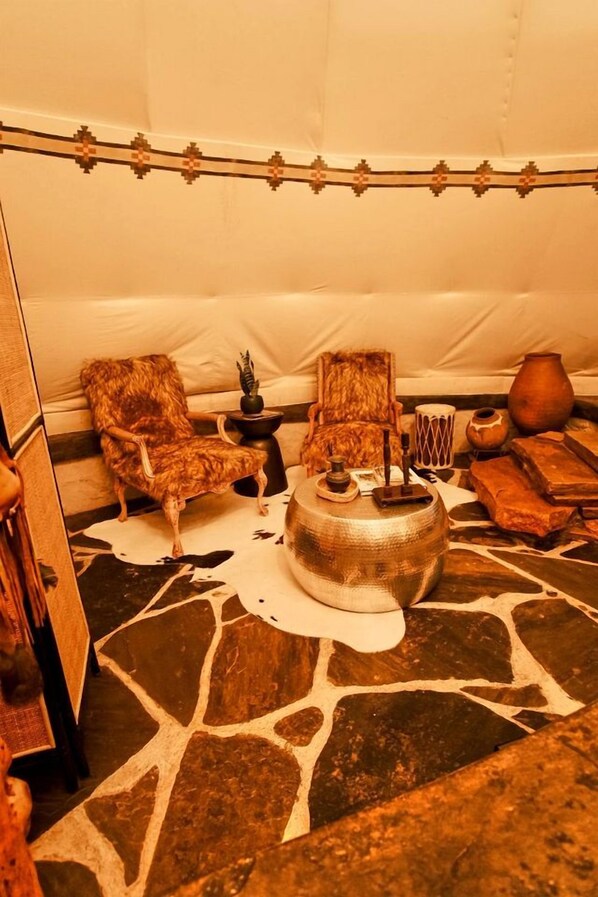 A tent interior with a stone floor, two chairs, a central table with a lamp, and various decorative items.