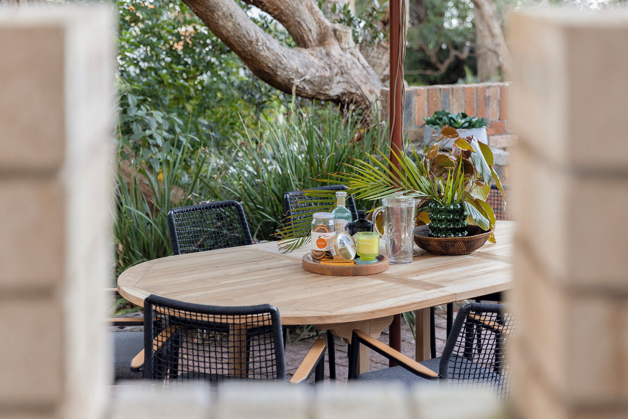 An outdoor dining area with a wooden table, black mesh chairs, and various items including a pineapple plant, a glass pitcher, and bottles.