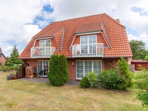 A two-story house with a red-tiled roof, a balcony on the upper floor, and a front yard with a small tree and shrubs. - Holiday Apartment With 2 Bedrooms (St. Peter-Ording)