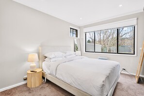 Ein Schlafzimmer mit Bett, Nachttisch, Lampe und einem Fenster mit Blick auf Bäume.