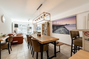 A modern living room with a dining area, a large window, and a beach view painting. - Apartment in Belgium Near the Harbor (Blankenberge)