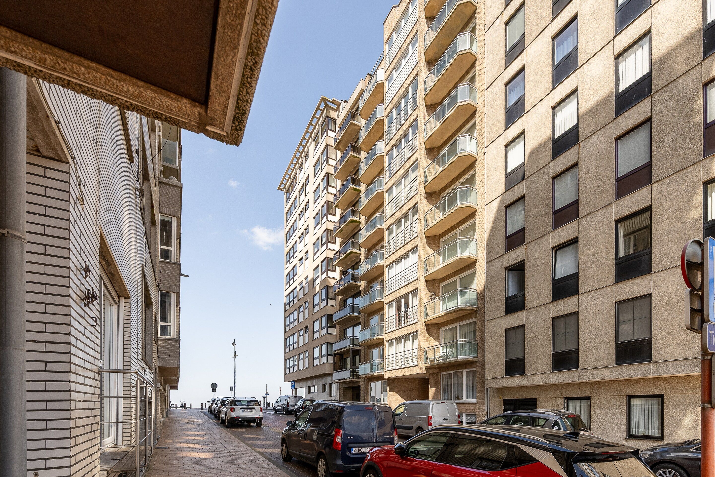 A multi-story residential building with balconies, parked cars, and a clear sky.
