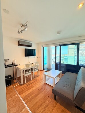Ein modernes Wohnzimmer mit einem grauen Sofa, einem Esstisch mit Stühlen, einem flachbildfernseher an der Wand und einem großen Fenster mit Blick auf die Stadt.