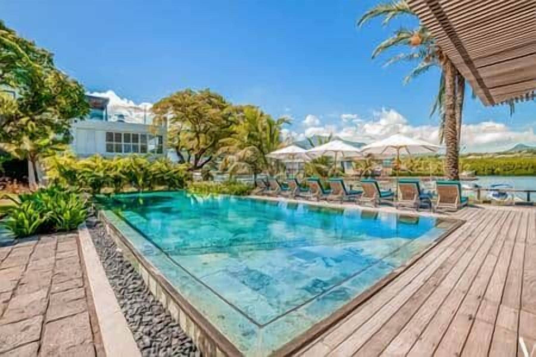 A swimming pool with lounge chairs and umbrellas, surrounded by tropical vegetation and a wooden deck.