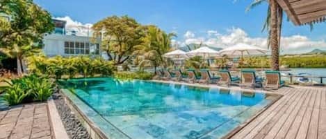 A swimming pool with lounge chairs and umbrellas, surrounded by tropical vegetation and a wooden deck.