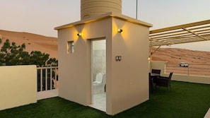 A small, enclosed outdoor area with a toilet, a chair, and a table, surrounded by artificial grass, with a view of a desert landscape.
