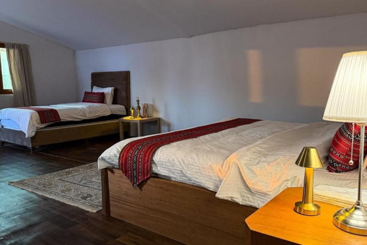 Ein Hotelzimmer mit zwei Betten, Holzboden, einem Nachttisch mit Lampe und einem Fenster mit Vorhängen.