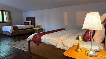 A hotel room with two beds, a wooden floor, a nightstand with a lamp, and a window with curtains.
