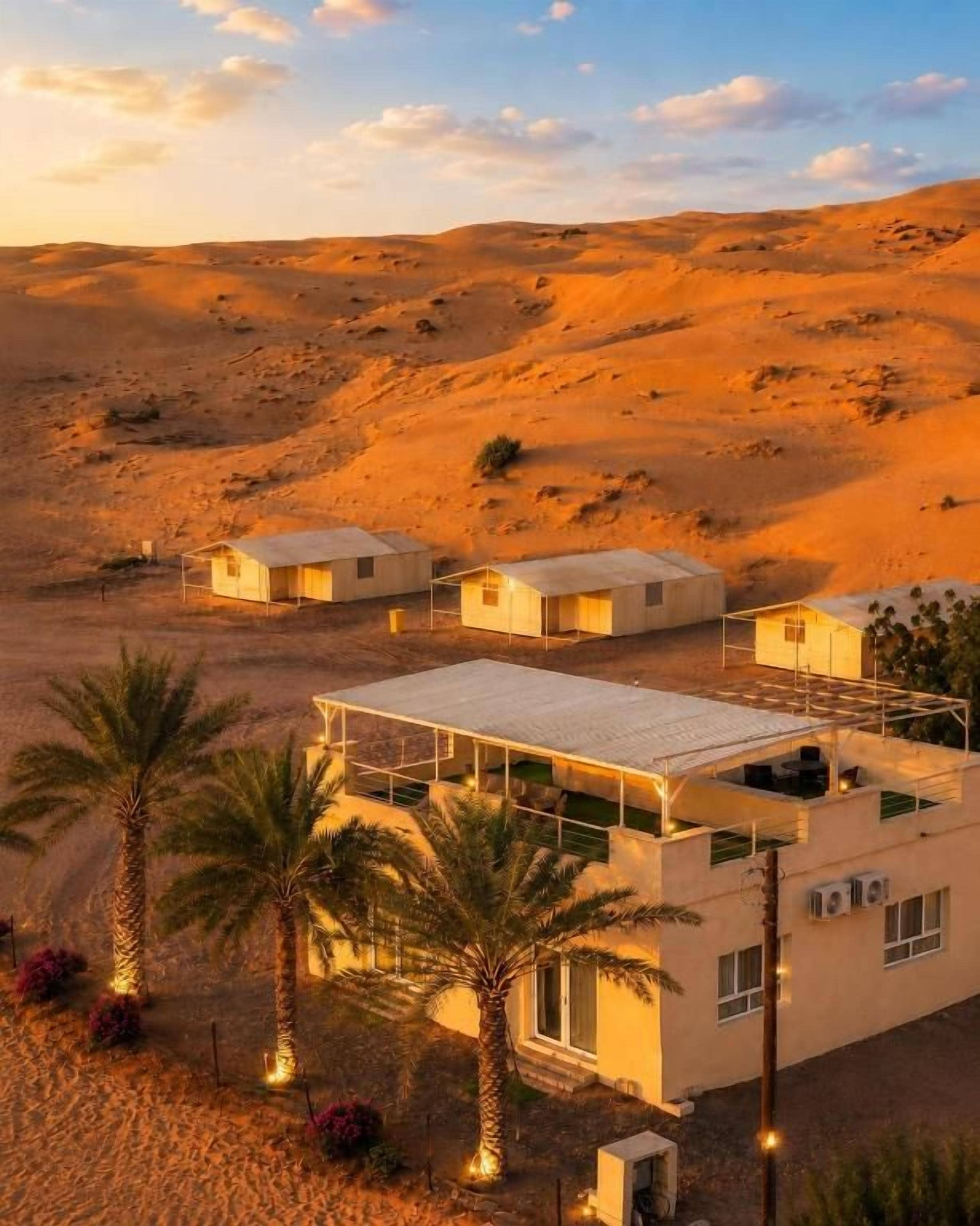 A desert landscape with a modern building featuring multiple rooms, palm trees, and a clear sky.