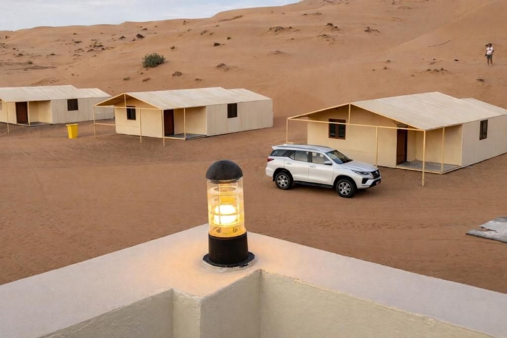 Paisaje desértico con varios edificios pequeños de una sola planta y techos planos, un vehículo blanco estacionado al frente y un poste de luz iluminado en primer plano.
