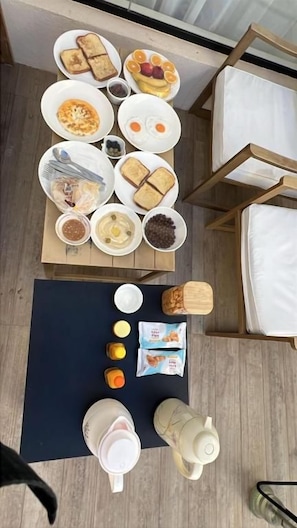 Ein Frühstücksbuffet mit verschiedenen Speisen und Getränken auf einem Tisch, einer Yogamatte und einem Bett mit weißen Laken.