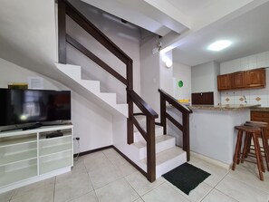 A kitchen with white cabinets, a wooden staircase, a black rug, and a television on a white stand.