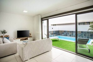 A modern living room with a flat-screen TV, two patterned sofas, and a large sliding glass door leading to a swimming pool. - Stylish Central Three Bed Duplex with Private Pool (Flic en Flac)