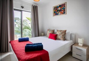 A hotel room with a bed, a nightstand, a lamp, and a window with curtains. - Stylish Central Three Bed Duplex with Private Pool (Flic en Flac)
