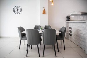 A modern dining area with a round table, six chairs, a wall clock, a hanging guitar, and a kitchen counter with a coffee machine. - Stylish Central Three Bed Duplex with Private Pool (Flic en Flac)
