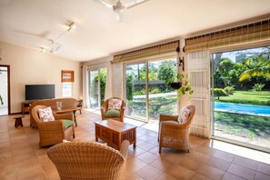 A spacious living room with wicker furniture, a flat-screen TV, and large sliding glass doors leading to a garden with a pool. - Cosy Bungalow with Pool and Beach Access (Black River)