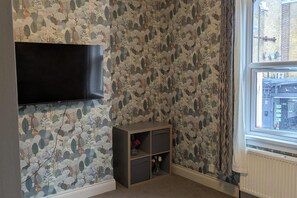 A room with a patterned wallpaper, a flat-screen TV mounted on the wall, a small wooden shelf with flowers, and a window with a view of buildings. - Central Stratford Gem (London)
