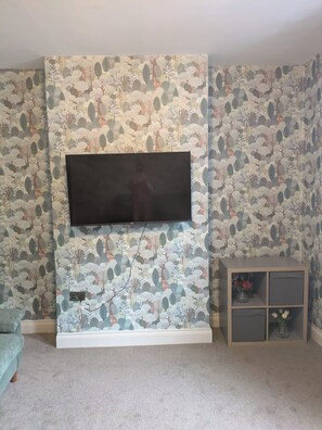 A room with a mounted flat-screen TV, patterned wallpaper, a carpeted floor, and a small wooden cabinet with flowers. - Central Stratford Gem (London)