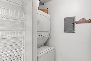 A laundry area with a stacked washer and dryer, a metal utility box, and a white metal shelf unit. - Pinnacle Port C1-403 (Panama City Beach)