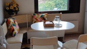 Una habitación pequeña y acogedora con una mesa redonda blanca, un sofá y dos sillones. Se ve un ventanal con vistas a zonas verdes.