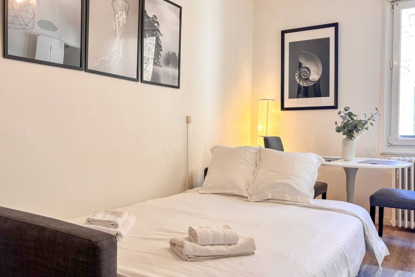 Une chambre d’hôtel avec un lit, un bureau et une chaise. Il y a des tableaux encadrés au mur, une lampe et une fenêtre.